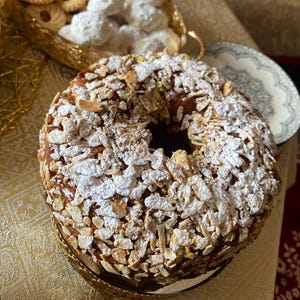 Può includere: Una torta rotonda con un buco al centro, ricoperta di mandorle a scaglie e zucchero a velo. La torta è su un piatto dorato. Altri pasticcini, tra cui biscotti e palle di neve, sono sullo sfondo.