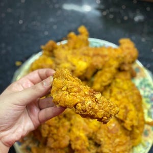 Może przedstawiać: Ręka trzyma złocistobrązowy, chrupiący smażony filet z kurczaka. Na tle talerz wypełniony podobnymi filetami. Jedzenie wydaje się świeżo ugotowane i gotowe do spożycia. Talerz ma kwiatowy wzór.