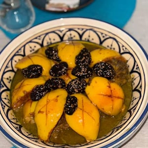 Könnte beinhalten: Ein traditionelles marokkanisches Tajine-Gericht mit gelben Früchten und dunklen, getrockneten Pflaumen. Das Gericht wird in einer dekorativen, blau-weißen Keramikschale serviert. Das Essen wird auf einer blauen Tischdecke präsentiert.