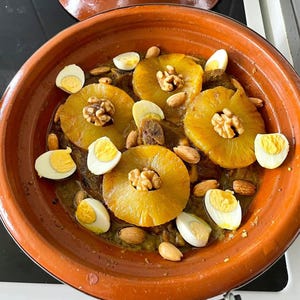 May include: A close-up shot of a Moroccan tagine dish in a terracotta pot. The dish features pineapple rings, walnuts, almonds, and halved hard-boiled eggs. The food is set against a dark background.