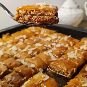 Op de afbeelding: Een close-up van baklava, een gelaagd gebak met noten en siroop. Een stuk baklava wordt op een vork gehouden, met de lagen en toppings van amandelschilfers. Het dessert ligt in een zwarte bakvorm.