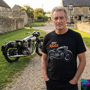May include: A man wearing a black t-shirt with the words "GHOST RIDER" and a motorcycle graphic. A vintage motorcycle is parked behind him on a gravel road. The background includes a stone wall and a building.