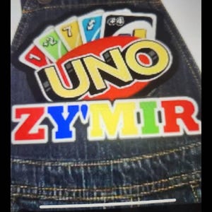 Może przedstawiać: Dżinsowe ogrodniczki z kolorową naszywką UNO. Naszywka przedstawia logo UNO i imię "ZYMIR" w wielokolorowych literach. Ogrodniczki mają regulowane paski i klasyczne detale dżinsowe.