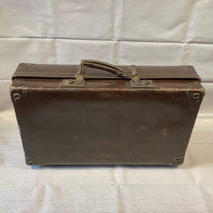 May include: A rectangular brown suitcase with a top handle, featuring reinforced metal corner protectors and two metal locking latches. The exterior surface shows signs of wear with visible scuffs and scratches across the dark brown material.