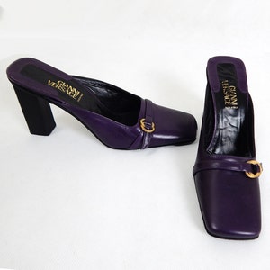 Peut inclure: Une paire de mules en cuir violet avec des bouts carrés et une boucle dorée. Les talons sont noirs et en forme de bloc. La marque "GIANNI VERSACE" est visible sur la semelle intérieure.