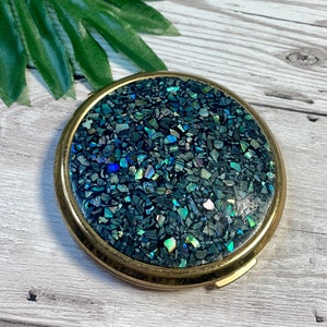 Könnte beinhalten: Ein runder Kompakt-Make-up-Spiegel mit einem goldfarbenen Metallrahmen. Die Oberseite zeigt ein Mosaik aus irisierenden, dunkelblauen und blauen Teilen. Der Kompaktspiegel ist geschlossen, mit einem kleinen Verschluss an der Seite. Heller Holzuntergrund.