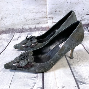 May include: A pair of dark gray suede high heels with pointed toes. Each shoe features a decorative flower with glitter accents and a metallic base. The heels are slender and the shoes are set against a light-colored wood background.