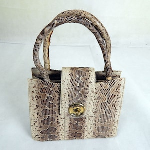 Könnte beinhalten: Eine kleine, quadratische Handtasche mit Schlangenmuster in Braun- und Cremetönen. Die Tasche hat einen goldfarbenen Verschluss und passende Griffe. Die Griffe sind gebogen und scheinen aus dem gleichen Schlangenleder-Material zu sein.