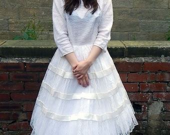 Puede incluir: Vestido blanco de encaje con falda escalonada y mangas largas. El vestido presenta detalles de cinta de raso y se combina con zapatos de tacón peep-toe color crema. Adecuado para una boda u ocasión especial.