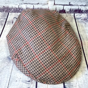 May include: A brown and black houndstooth pattern flat cap with red accent lines. The hat is made of a textured fabric and is designed in a classic style. The cap is a traditional accessory.