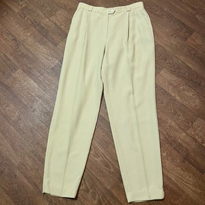 May include: Pale yellow trousers featuring a high-waisted design with a single button closure at the center. These pants include vertical pleats extending from the waistband down the front of each leg, creating a tailored look with straight legs.