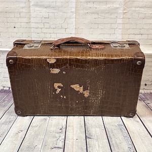 May include: A vintage brown suitcase with a textured, crocodile-effect pattern. The suitcase features a leather handle and metal corner protectors. The finish shows signs of wear, adding to its antique charm. The rectangular suitcase has metal clasps.