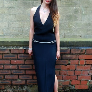 May include: A black halter-neck dress with a V-neckline and a silver chain belt. The dress is floor-length with a side slit. The person is wearing a silver necklace, bracelet, and white heels.