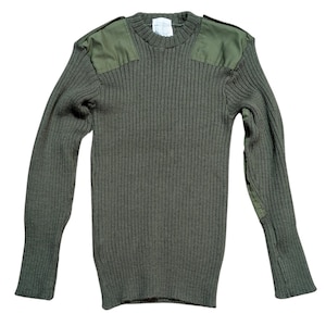 Puede incluir: Un suéter de manga larga color verde oliva con textura de punto acanalado, cuello redondo y parches de tela reforzados en los hombros y los codos. Una etiqueta rectangular blanca con texto impreso es visible en el área del cuello.