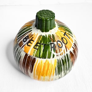 May include: A ceramic jar with a lid, painted with vertical stripes in shades of green, yellow, brown, and white. The jar is labeled "BEET ROOT" in black lettering. The lid is green and shaped like a stem.