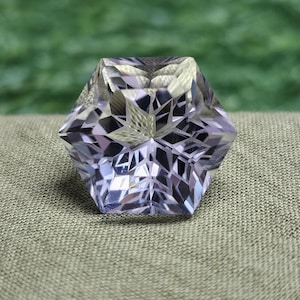 Puede incluir: Una gema facetada de color púrpura claro con un complejo patrón interior en forma de estrella. La gema tiene forma hexagonal y se encuentra sobre una tela gris texturizada, con un fondo verde borroso.
