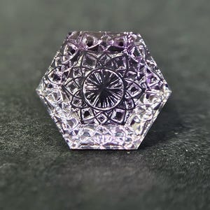 Peut inclure: Une pierre précieuse hexagonale violette translucide présentant un motif géométrique détaillé et complexe gravé sur sa surface. La pierre affiche un design à facettes avec un motif central en étoile entouré de formes florales symétriques.