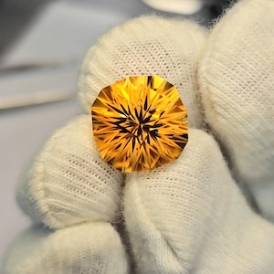 Puede incluir: Una gema facetada de color amarillo dorado con un patrón de estrella. La gema está sujeta por una mano enguantada, mostrando sus intrincados cortes y su color brillante. La forma de la gema es un corte cojín modificado.