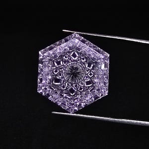 May include: A hexagonal, purple gemstone with intricate facets is held by tweezers against a black backdrop. The gem has a symmetrical design with a central starburst pattern and detailed facets.