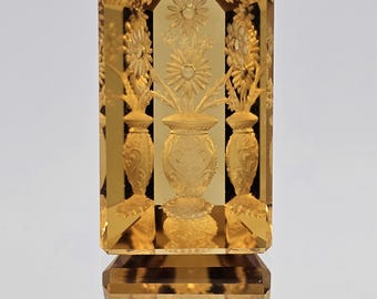 May include: A golden-colored, rectangular glass object with a floral design. The object features a central vase with flowers and two side vases. The top is arched and also has a floral pattern. The base is a solid, rectangular shape.