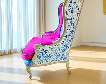 May include: A tall, ornate armchair with a high back. The chair features a silver-painted wooden frame, a bright pink seat and back, and a white fabric back with blue splatter paint. The edges are trimmed with small, silver studs.