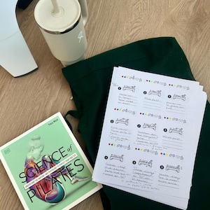 May include: A book titled "Science of Pilates" with a green cover and a woman illustration. A white mug with a straw, a dark green garment, and a sheet of paper with handwritten notes and diagrams are also visible.