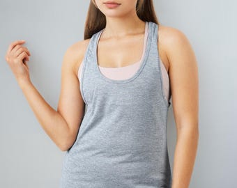 May include: A gray tank top with a racerback design. The tank top is layered over a light pink sports bra. The model is wearing light pink leggings. The background is a solid gray.