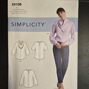 May include: A Simplicity sewing pattern package featuring a woman wearing a lilac-colored blouse and blue pants. The package includes illustrations of different blouse styles. The text on the package reads "S9108" and "SIMPLICITY".