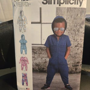 May include: A Simplicity S9486 sewing pattern envelope for a hooded romper in sizes 1/2-4. The front displays four variations of the garment, including a blue short-sleeve hooded romper with front pleats and pockets, alongside three other printed designs.