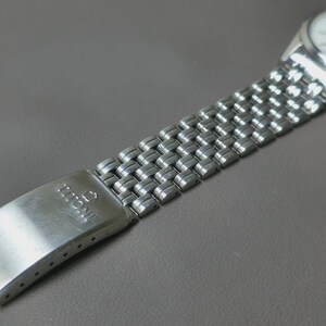 May include: Close-up of a silver-toned wristwatch with a white face and a stainless steel band. The band features a woven design. The clasp is marked with the brand name "TITONI". The watch is set against a gray background.