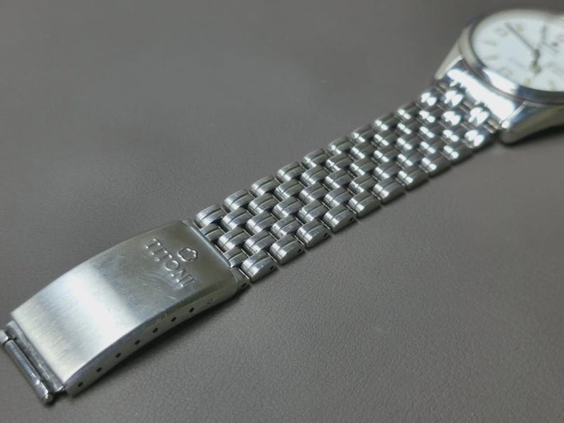 May include: Close-up of a silver-toned wristwatch with a white face and a stainless steel band. The band features a woven design. The clasp is marked with the brand name "TITONI". The watch is set against a gray background.