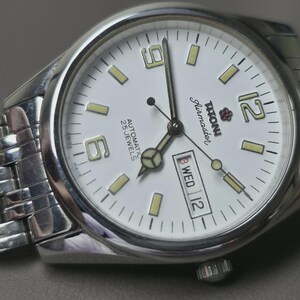 May include: Silver-toned wristwatch with a white face, featuring black hands and markers. The watch face displays the day and date, with the brand name "Titoni Airmaster". The watch has a silver band and is set against a neutral background.