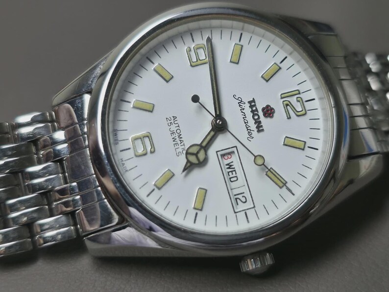May include: Silver-toned wristwatch with a white face, featuring black hands and markers. The watch face displays the day and date, with the brand name "Titoni Airmaster". The watch has a silver band and is set against a neutral background.
