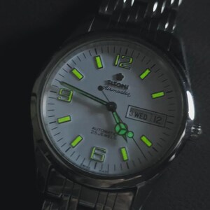 May include: Silver-toned wristwatch with a light gray face. The watch features luminous green hour markers and hands, with the day and date displayed at the 3 o'clock position. The watch is labeled "Tilton Airmaster" and has a stainless steel band.