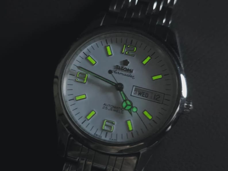 May include: Silver-toned wristwatch with a light gray face. The watch features luminous green hour markers and hands, with the day and date displayed at the 3 o'clock position. The watch is labeled "Tilton Airmaster" and has a stainless steel band.