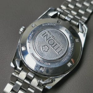 May include: Close-up of the back of a silver-toned Tissot watch. The watch features a stainless steel band and a circular back with the brand name and "of Switzerland" engraved. The watch is labeled "Automatic" and "Water Resistant".