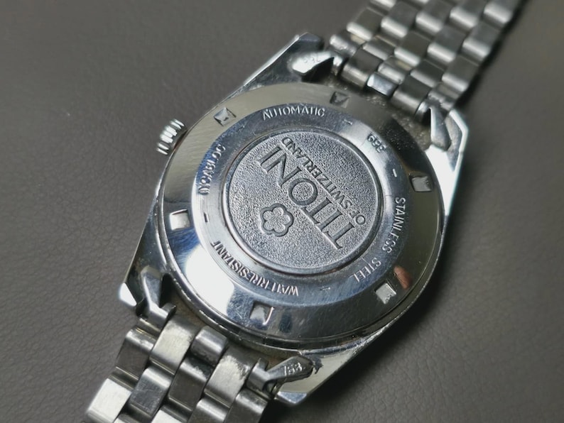 May include: Close-up of the back of a silver-toned Tissot watch. The watch features a stainless steel band and a circular back with the brand name and "of Switzerland" engraved. The watch is labeled "Automatic" and "Water Resistant".