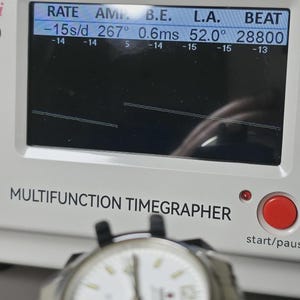 May include: A white-faced wristwatch is positioned in front of a multifunction timegrapher. The timegrapher displays readings for RATE, AMP, B.E., L.A., and BEAT. A red start/pause button is visible on the timegrapher.