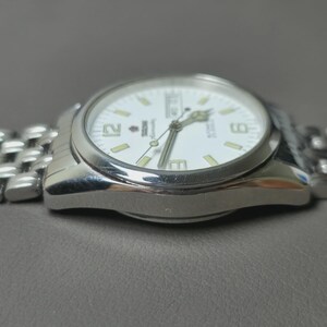 May include: Silver-toned wristwatch with a white face, featuring black numerals and hands. The watch has a stainless steel band and a date display. The watch is shown on a gray surface.