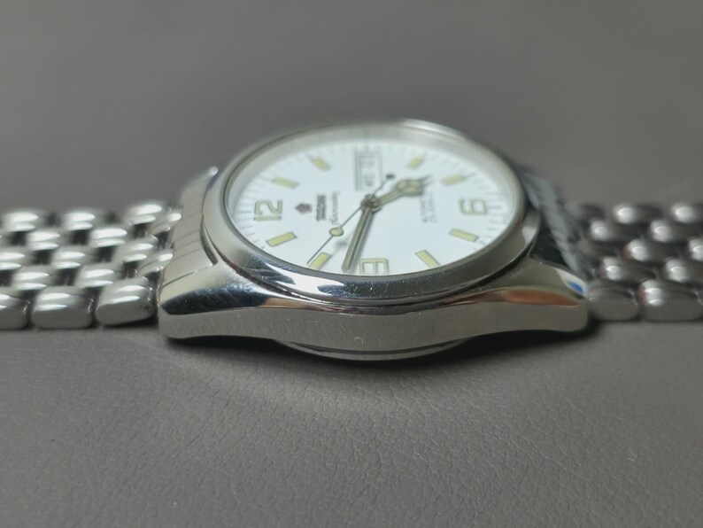 May include: Silver-toned wristwatch with a white face, featuring black numerals and hands. The watch has a stainless steel band and a date display. The watch is shown on a gray surface.