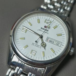 May include: Silver-toned wristwatch with a white face. The watch features a day and date display, luminous markers, and the text "Titoni Airmaster Automatic 25 Jewels". The watch has a silver band.