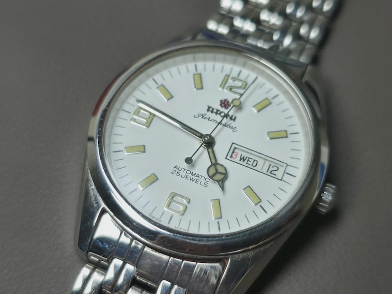 May include: Silver-toned wristwatch with a white face. The watch features a day and date display, luminous markers, and the text "Titoni Airmaster Automatic 25 Jewels". The watch has a silver band.