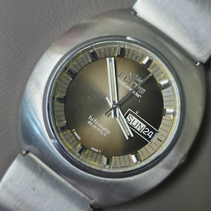 May include: A vintage Fortis hi-fi-matic spacelander watch with a silver case and strap. The watch face is a gradient brown colour with silver hands and markers. The day and date are displayed at the 3 o'clock position. The watch reads "SUN24".