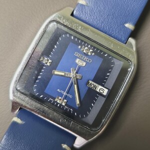 May include: A vintage Seiko 5 automatic watch with a blue dial and a silver-tone case. The watch features a blue leather strap and displays the day and date. The watch face has silver-tone hands and markers.