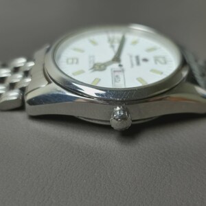 May include: A silver wristwatch with a white face and a silver band. The watch face displays the time with black numerals and hands, and a day-date window. The watch is on a gray surface.