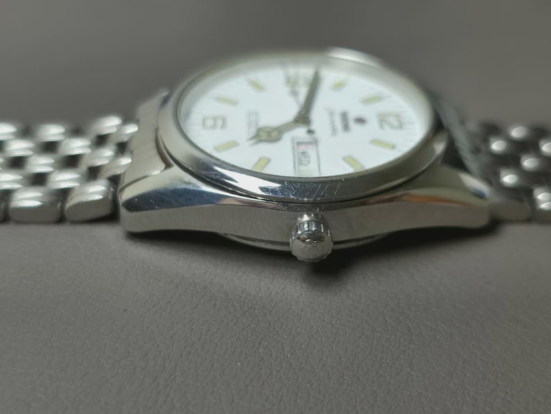 May include: A silver wristwatch with a white face and a silver band. The watch face displays the time with black numerals and hands, and a day-date window. The watch is on a gray surface.