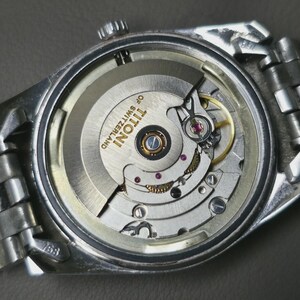 May include: Close-up of a silver wristwatch with the back removed, revealing the intricate mechanical movement. The watch mechanism includes gears, jewels, and the text "TITONI OF SWITZERLAND". The watch has a silver metal band.