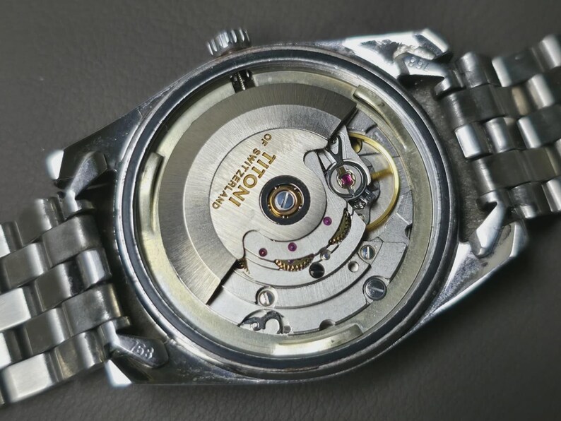 May include: Close-up of a silver wristwatch with the back removed, revealing the intricate mechanical movement. The watch mechanism includes gears, jewels, and the text "TITONI OF SWITZERLAND". The watch has a silver metal band.