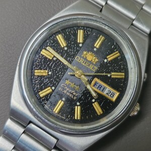 May include: Close-up of a silver-toned Orient Crystal watch with a black dial and gold-colored hands and markers. The watch face displays the day and date, with the day reading "FRI 28". The watch has a silver-toned metal band.