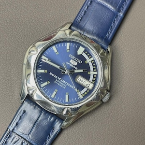 Puede incluir: Reloj de pulsera automático Seiko 5 Sports con esfera azul y correa de cuero azul. El reloj tiene caja plateada, visualización de día y fecha y es resistente al agua hasta 100m. Muestra el día 'JUE 4'.